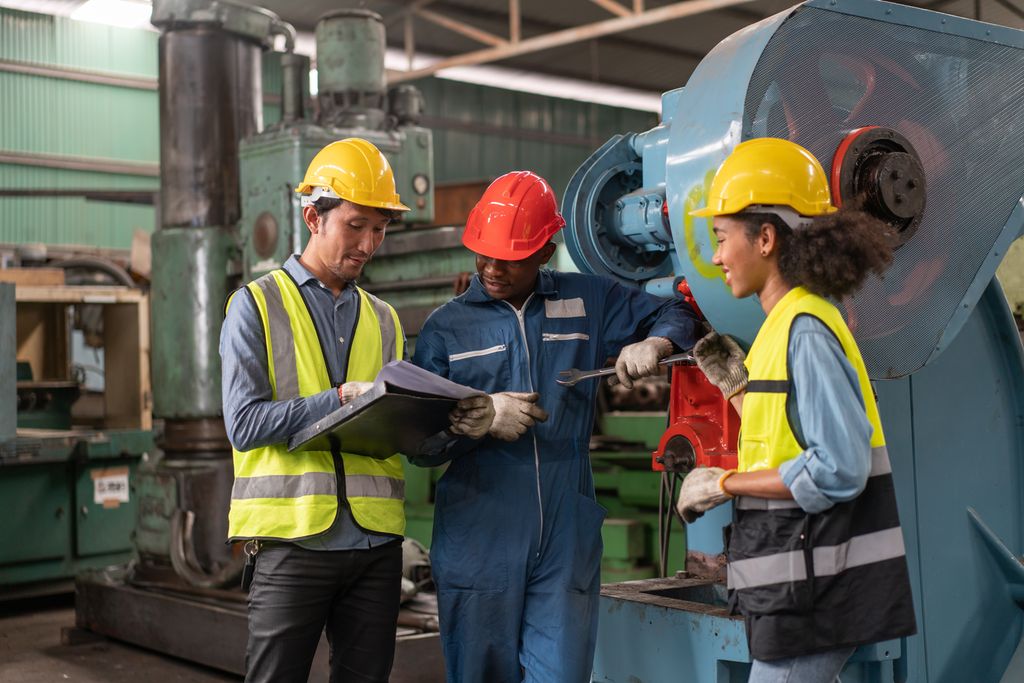 Tre ingeniører med sikkerhetsvest og hjelm studerer spesifikasjonsdokumenter for reparasjon og vedlikehold på en fabrikk. Foto.