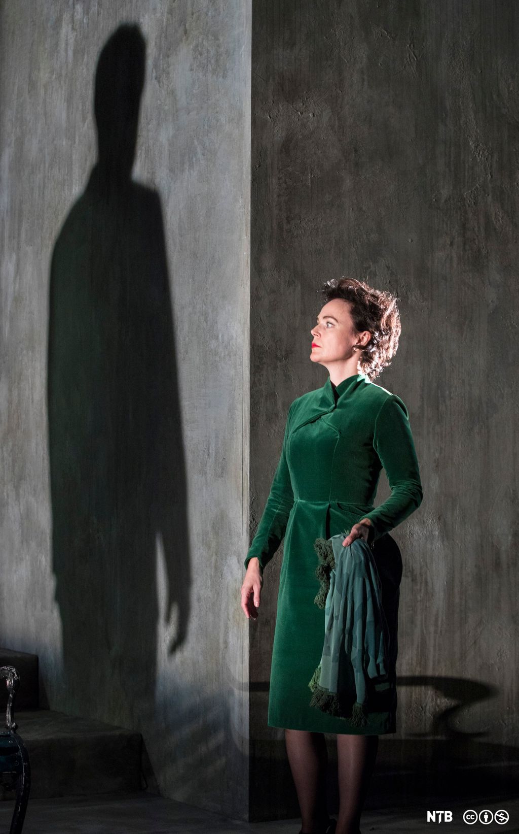 Fru Alving, kledd i grønt, står inntil en  vegg og ser på skyggen til pastor Manders.