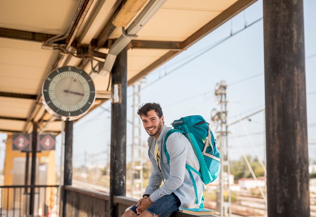 Smilende ung mann med ryggsekk på perrongen til en togstasjon. I bakgrunnen skimtes togskinner og elektriske ledninger. Foto.