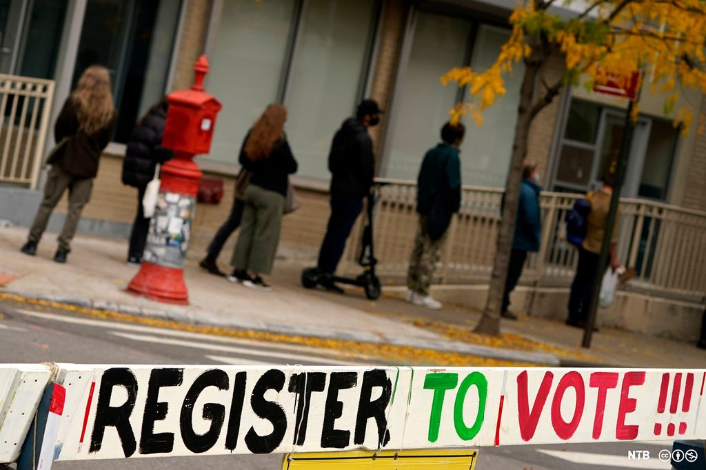 A sign urging people to register to vote is displayed near a line of people at a voting site in New York during the 2020 election. Photo.