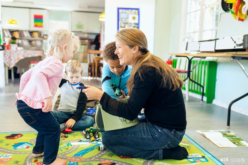 En voksen sitter på gulvet sammen med flere barn. Hun smiler og gir en lekebil til et barn. Foto. 