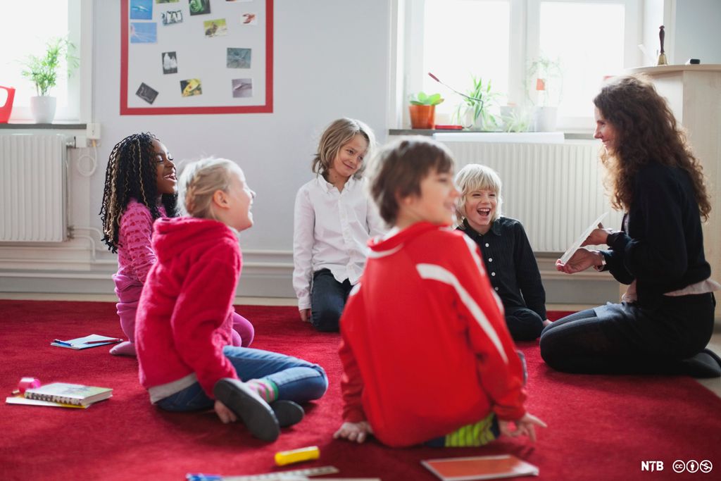 Ei gruppe barn sitter i en ring på gulvet sammen med en voksen. Den voksne viser barna noe. Alle smiler og ler. Foto.