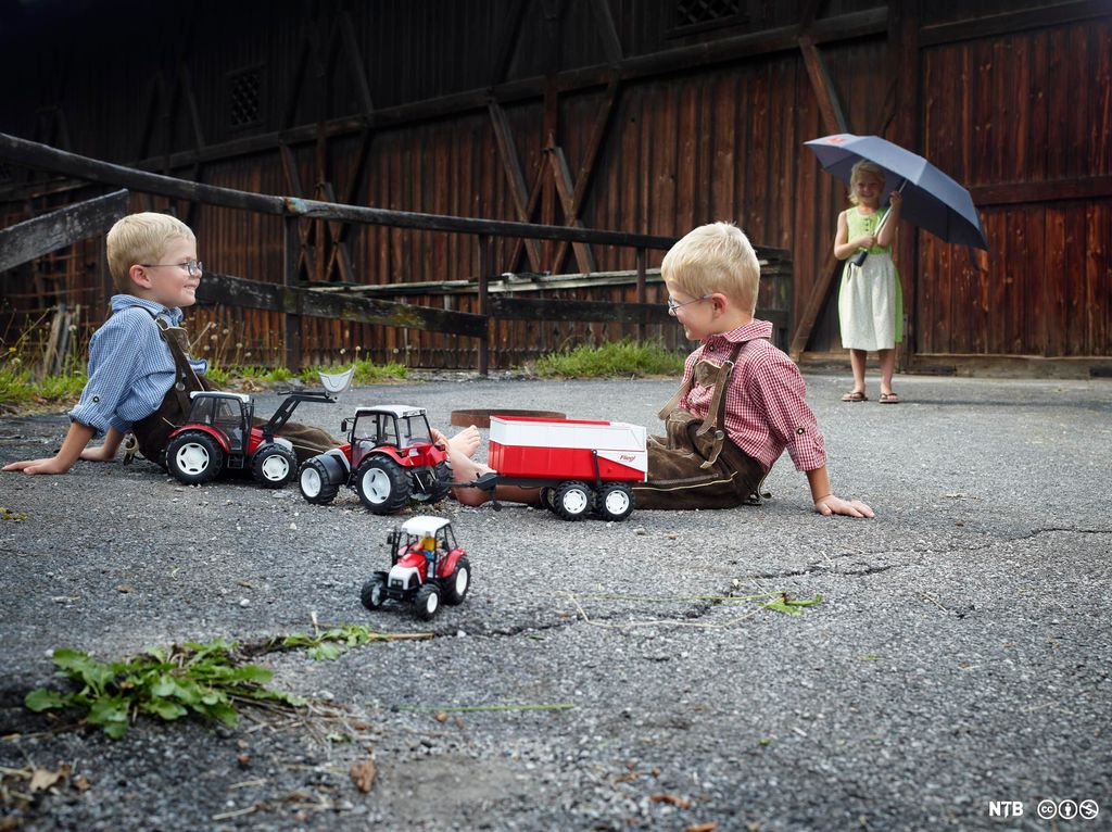 To gutter sitter på bakken ved siden av tre leketraktorer. Ei jente står litt lenger unna med en paraply. Det regner ikke, og barna har sommerklær. Foto.