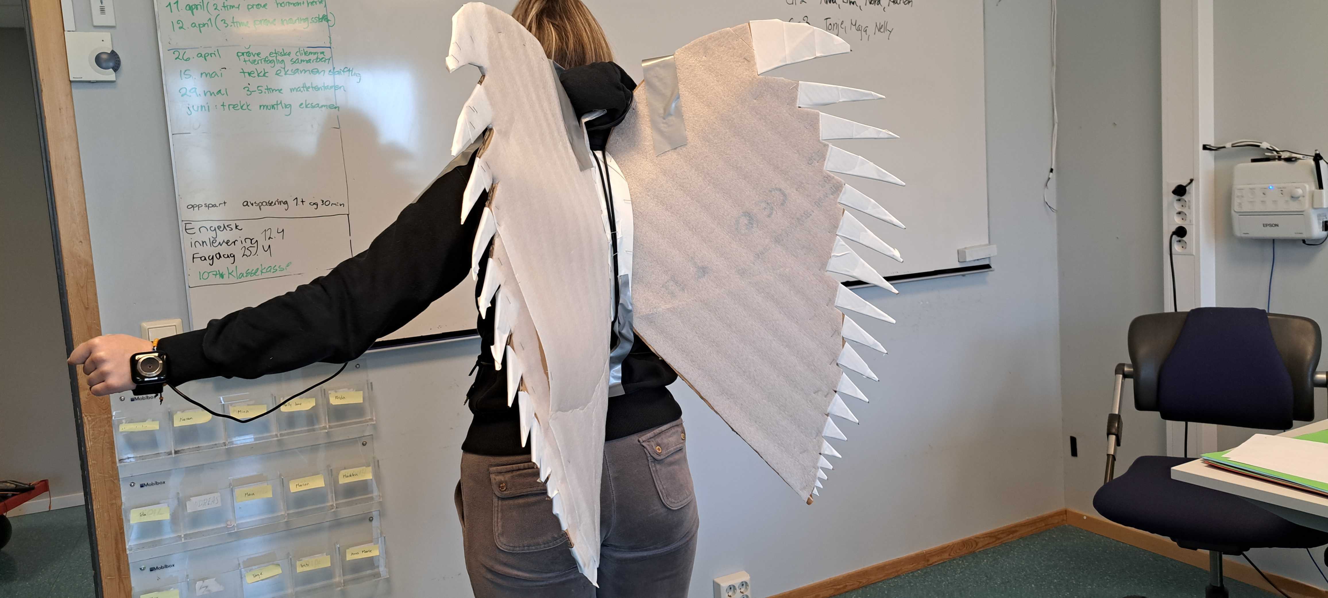 Person med vinger av papp på ryggen. Vingene er koplet til et armbåndsur med ledninger. Foto.