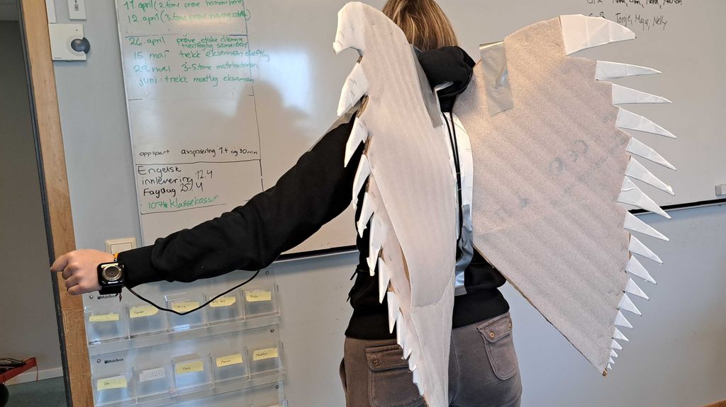 Person med vinger av papp på ryggen. Vingene er koplet til et armbåndsur med ledninger. Foto.