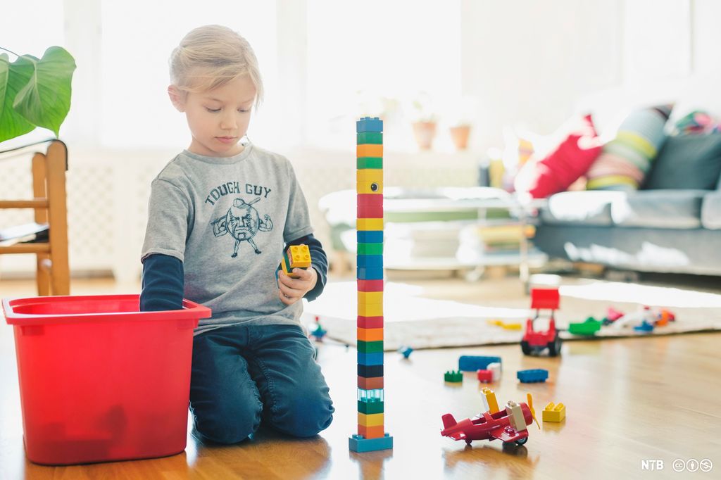 Jente som sitter og bygger tårn med store legoklosser. Ved siden av tårnet står et legofly, og flere andre klosser ligger utover gulvet. Foto.