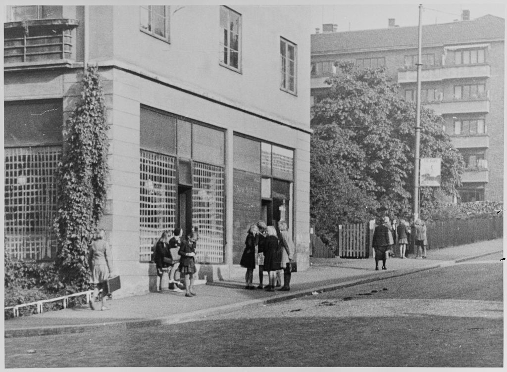 Barn utenfor en bygning med gitter foran vinduene i første etasje. I bakgrunnen er det busker, trær og ei boligblokk. Foto. 
