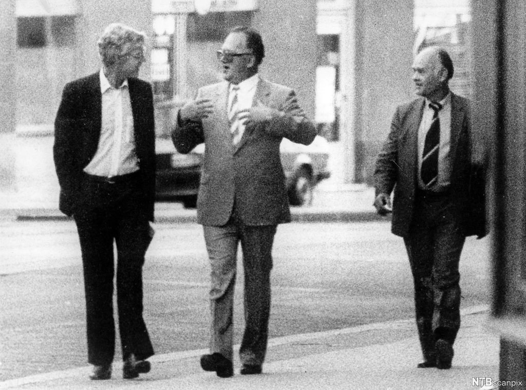 Tre menn kledde i dress går og samtaler med kvarandre i ei gate. Svart-kvitt-foto.