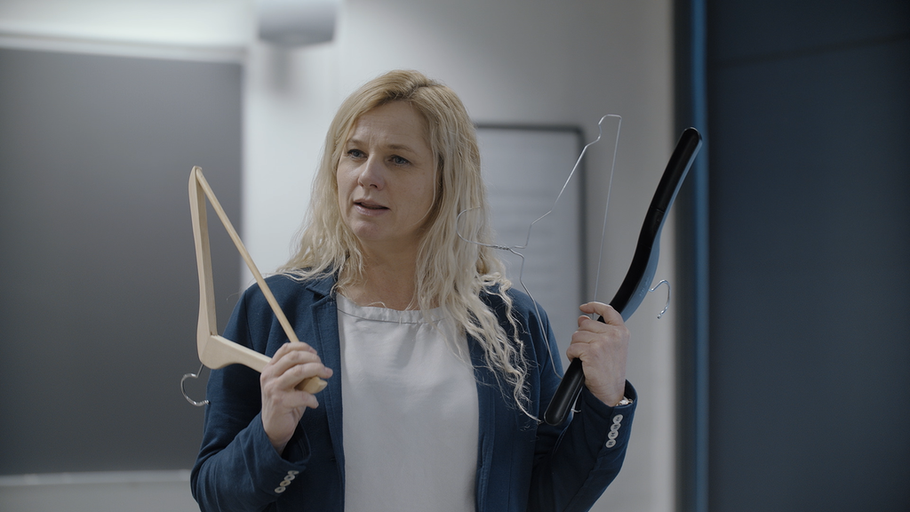 Lærer med blå jakke og blondt hår står foran tavla i et klasserom og holder fram tre ulike kleshengere: en i plast, en i metall og en i tre. Foto.