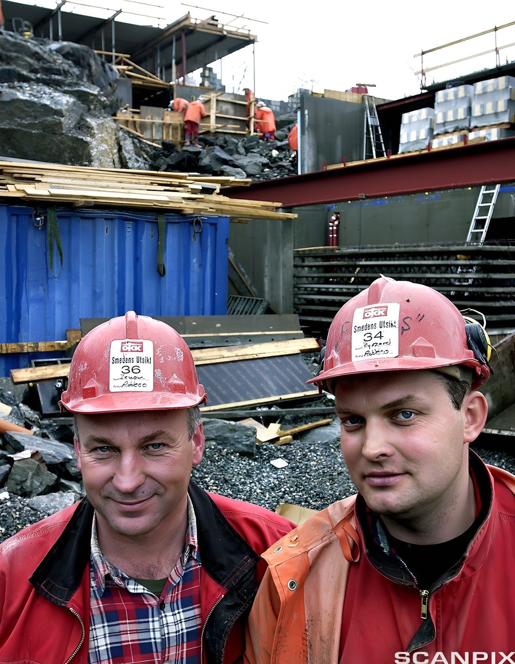 To smilande arbeidarar i raude arbeidsklede og med hjelm står på ein byggeplass. Foto. 