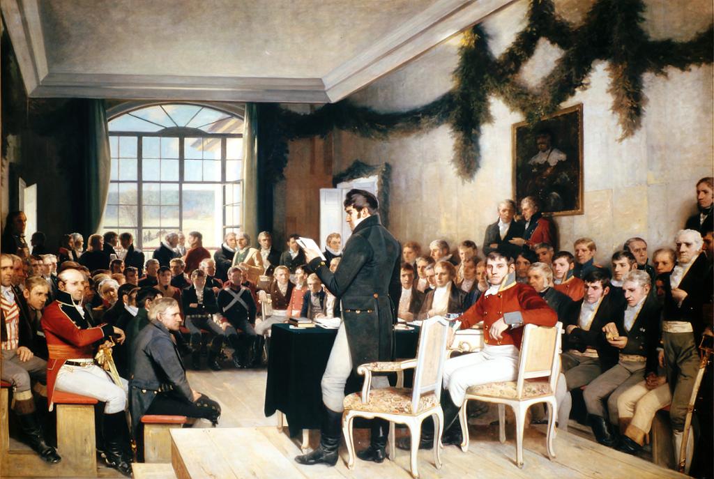 Det kjende måleriet av riksforsamlinga på Eidsvoll 1814. Ei forsamling menn sit på benker i ein sal. Dei fleste har knebukser og lange jakker. Ved eit skrivebord sit ein mann, ein annan står ved bordet og les frå eit ark. 