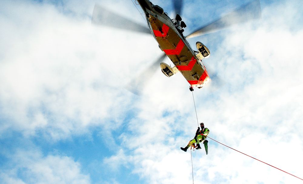 Mann som henger i stropp under et helekopter, og dras mot høyre av en vaier. Fotografi.