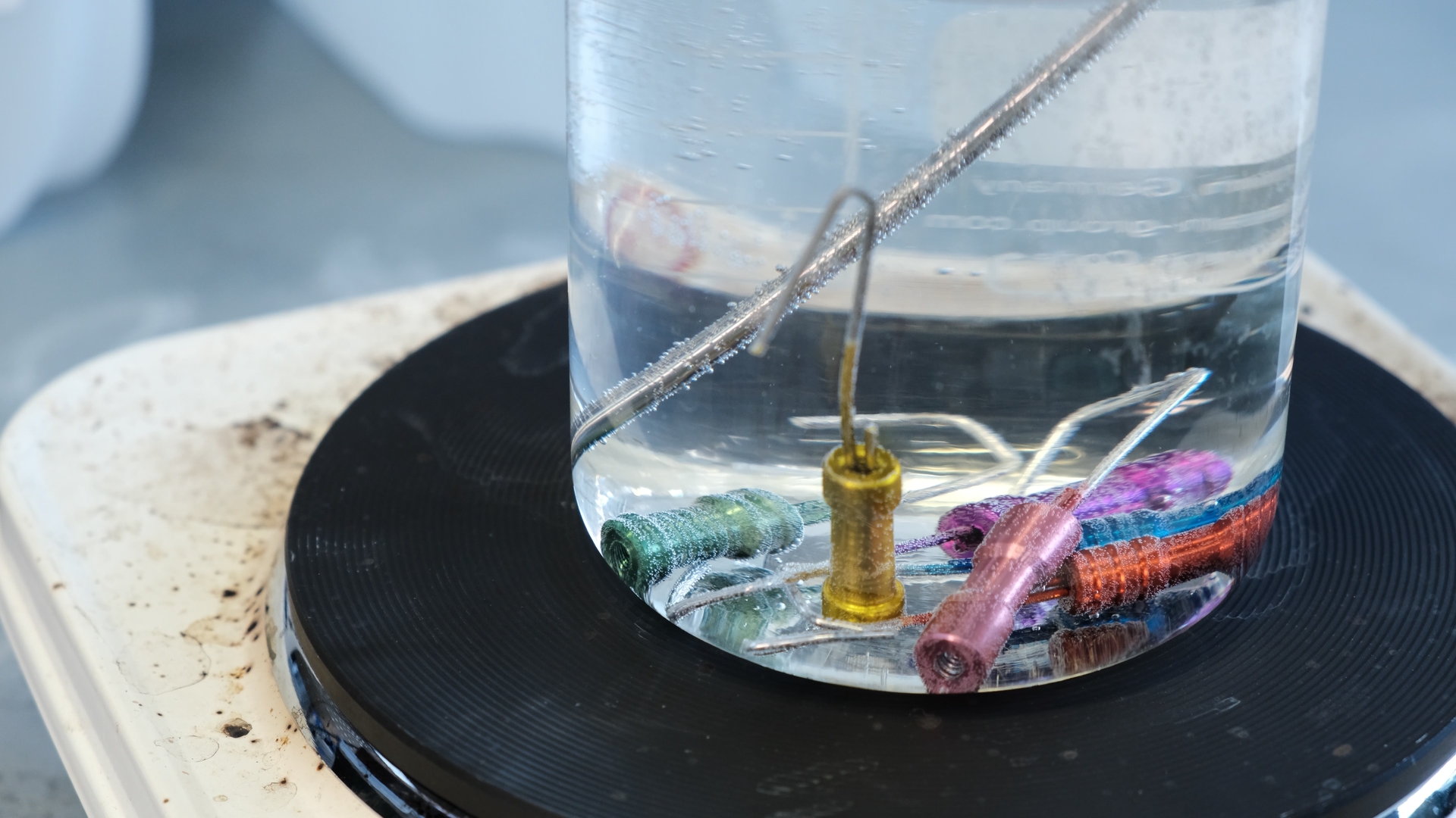 Metalldeler med ulik farge som ligger i en væskefylt glassbeholder som står på ei kokeplate. Foto.