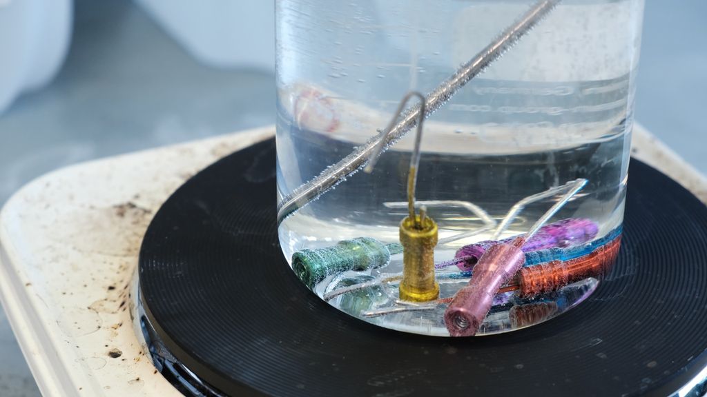 Metalldeler med ulik farge ligger i en væskefylt glassbeholder som står på ei kokeplate. Foto.