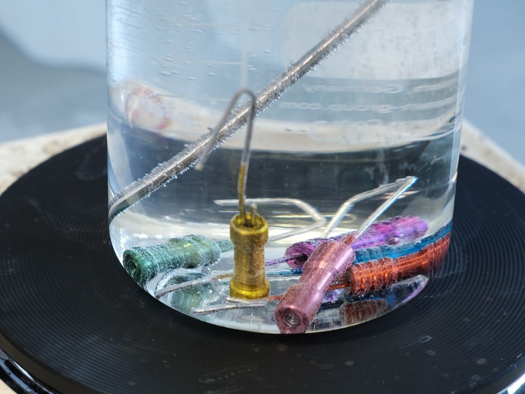 Metalldeler med ulik farge som ligger i en væskefylt glassbeholder som står på ei kokeplate. Foto.