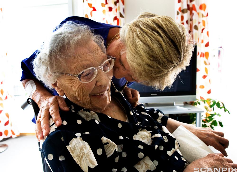 Omsorgsarbeider står bak ei eldre dame som sitter i en stol. Omsorgsarbeideren har hendene på skuldrene til dama og bøyer seg mot øret hennes for å si noe. Foto. Foto.