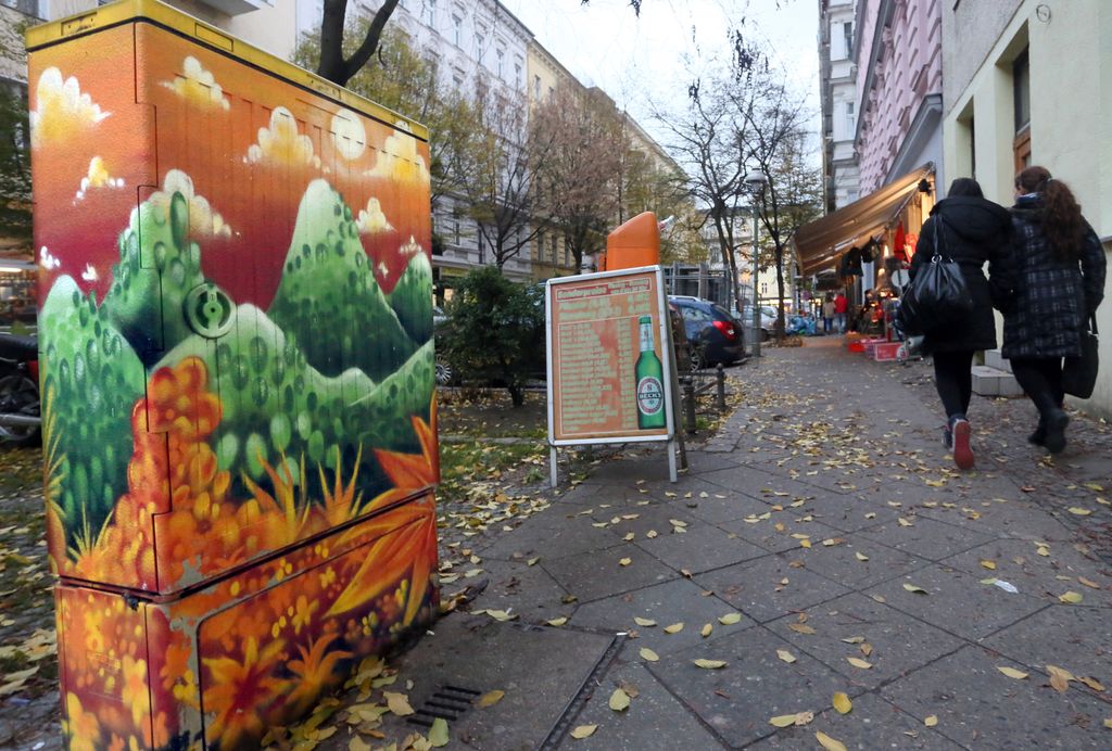 Ei gate i Berlin. Det er høst,  himmelen er overskya og det ligger gult løv på bakken. I forgrunnen et lite el-anlegg med grønn og oransje graffiti på. Foto.