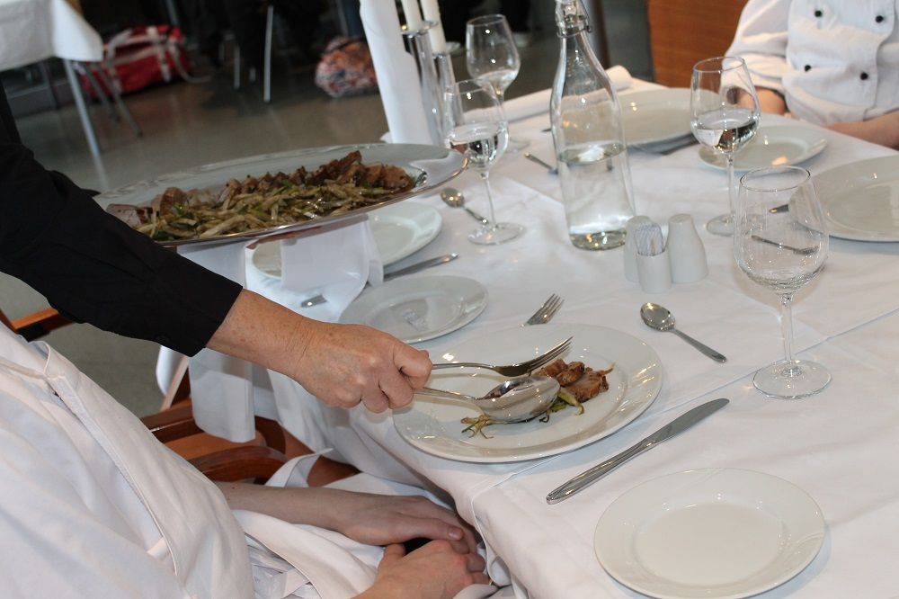 En servitør legger mat på gjestens tallerken fra gjestens vestre side. Foto.