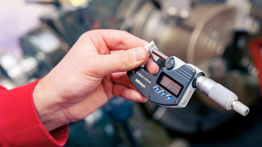 Ei hånd holder et måleverktøy som måler en liten, hvit del. Displayet på verktøyet viser 2,000. Foto.