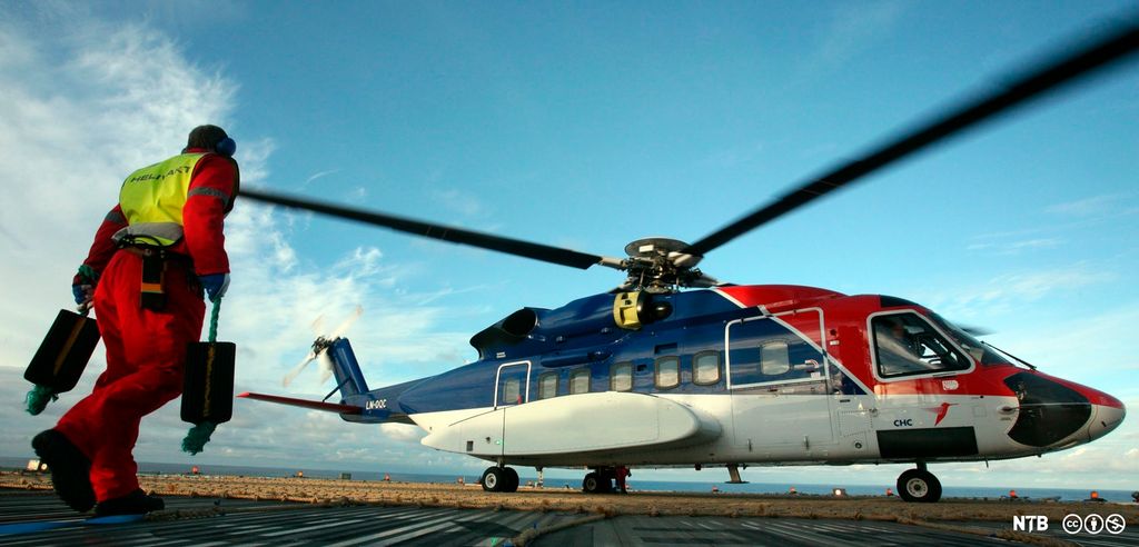 Ein person i kjeledress og med refleksvest og øyreklokker er i på veg mot eit Sikorsky S92-helikopter. Personen har ein hjulkloss i kvar hand. Foto.