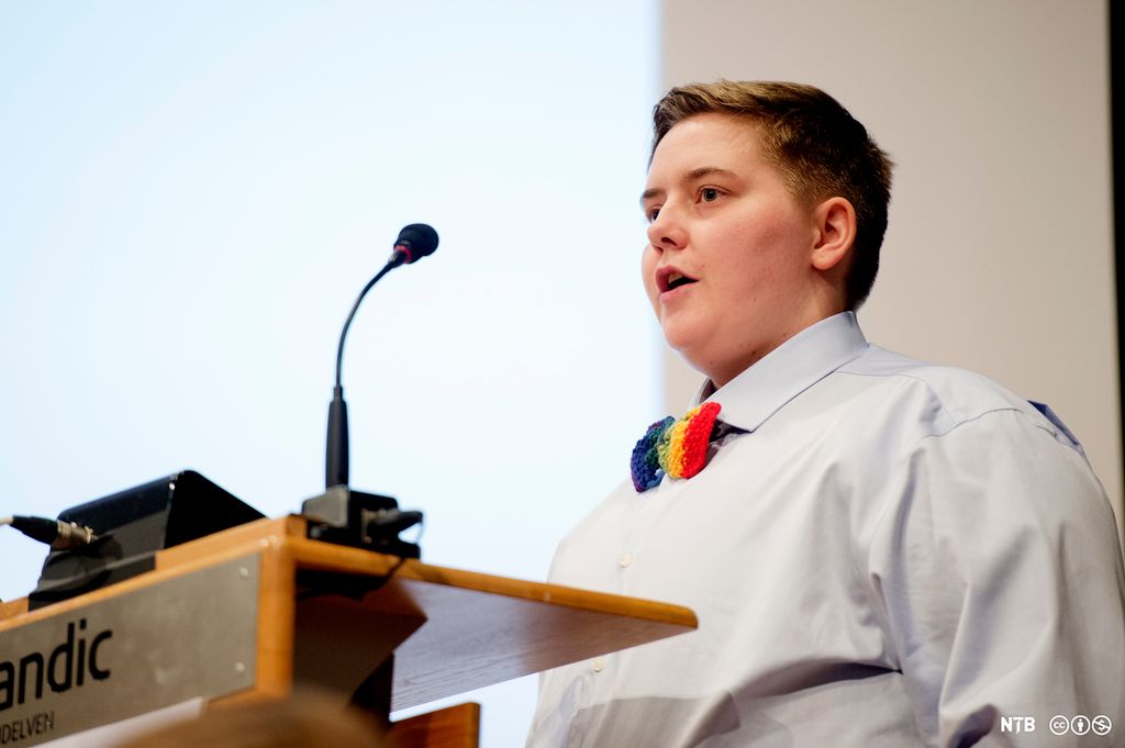 En person med kortklipt hår, kledd i skjorte og med regnbuefarget sløyfe, står på talerstolen. Foto.