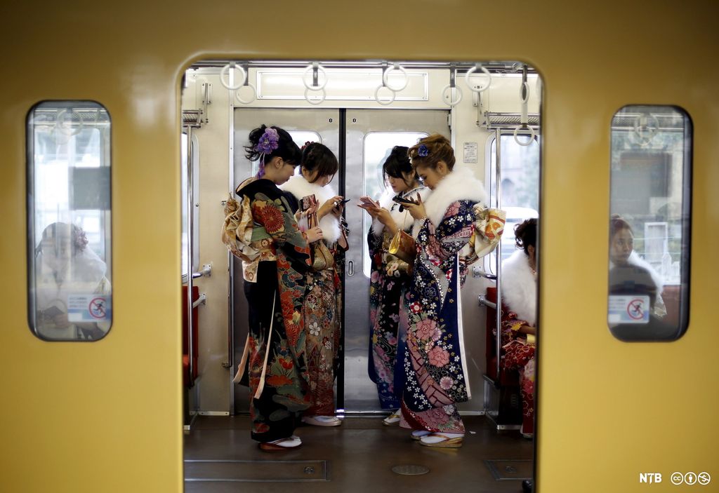 Train doors are open. We see four women in traditional Japanese clothes. They are looking at their phones. Photo.