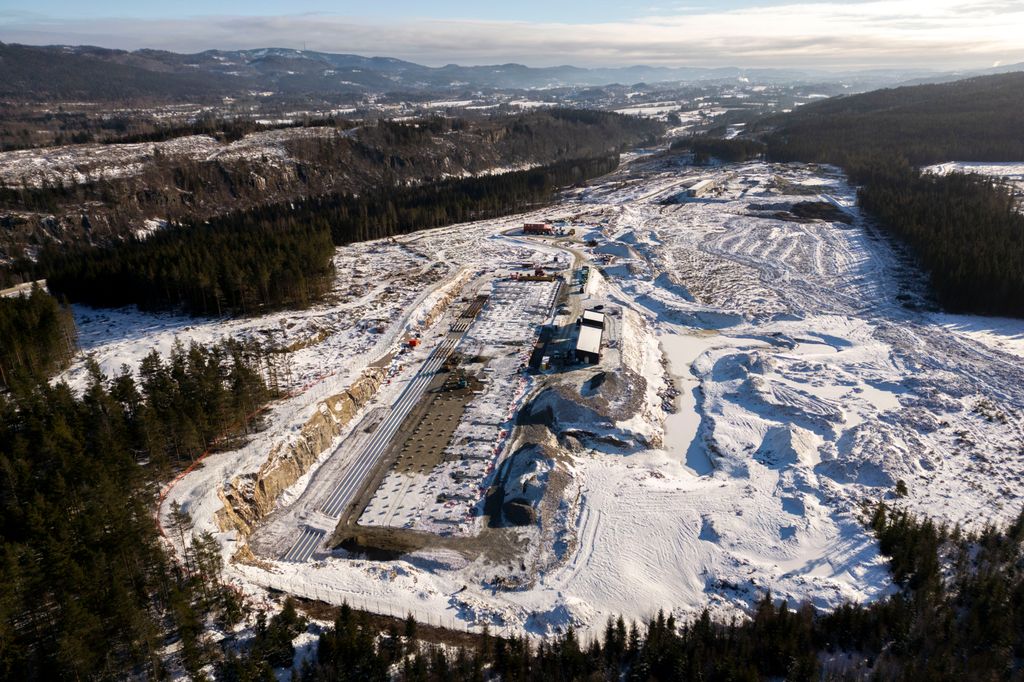 Stort bygg under konstruksjon. Jorder og skog omgir byggeplassen. Foto.