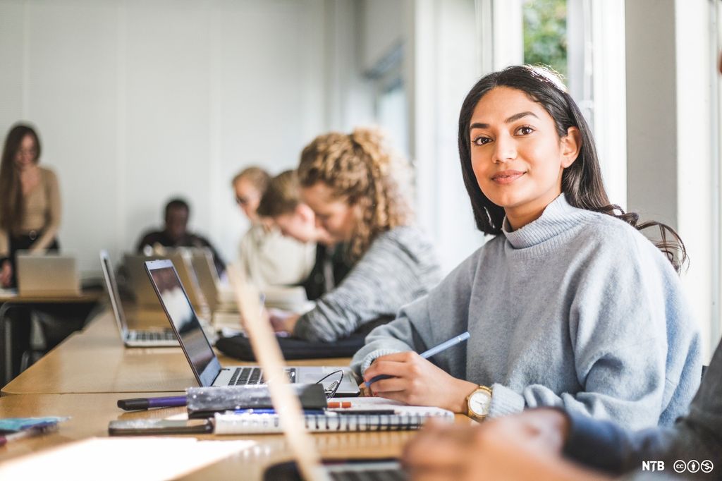 En ung kvinne sitter foran pc-en sin i et klasserom. I bakgrunnen sitter flere personer med pc på pultene sine. Foto. 