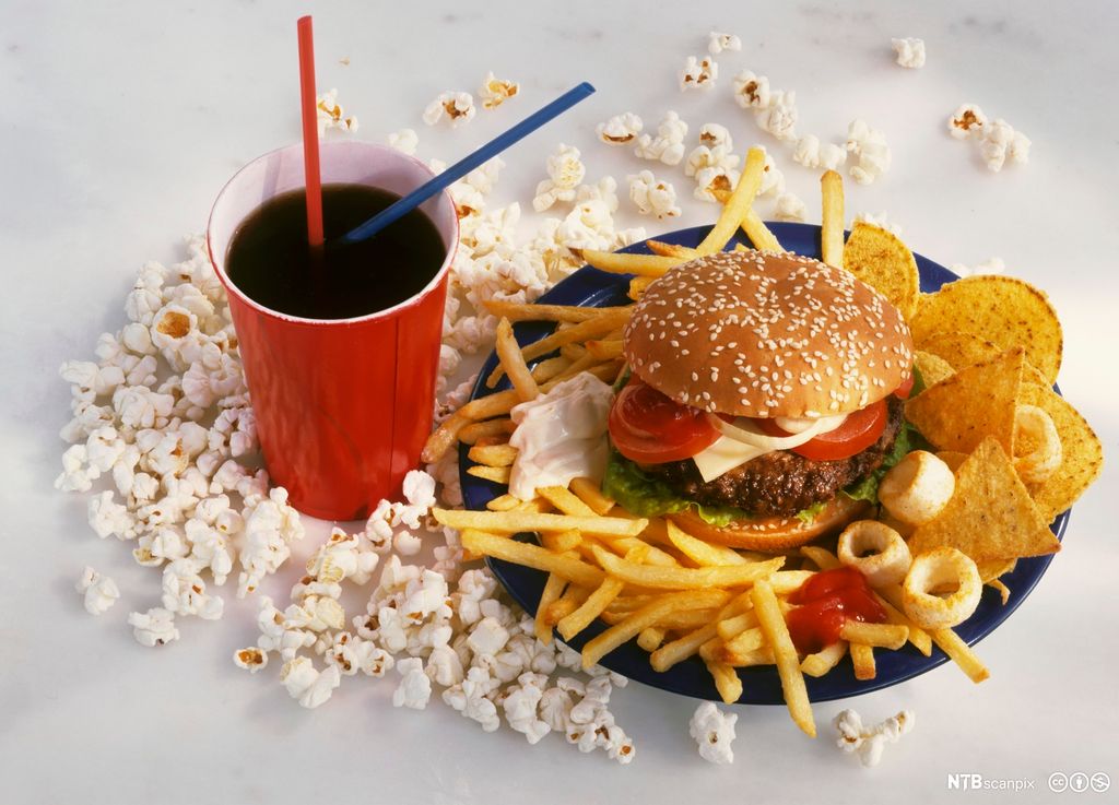 Pappbeger med brus og en tallerken med hamburger med pommes strips og tortillachips. Det ligger popkorn rundt. Foto.
