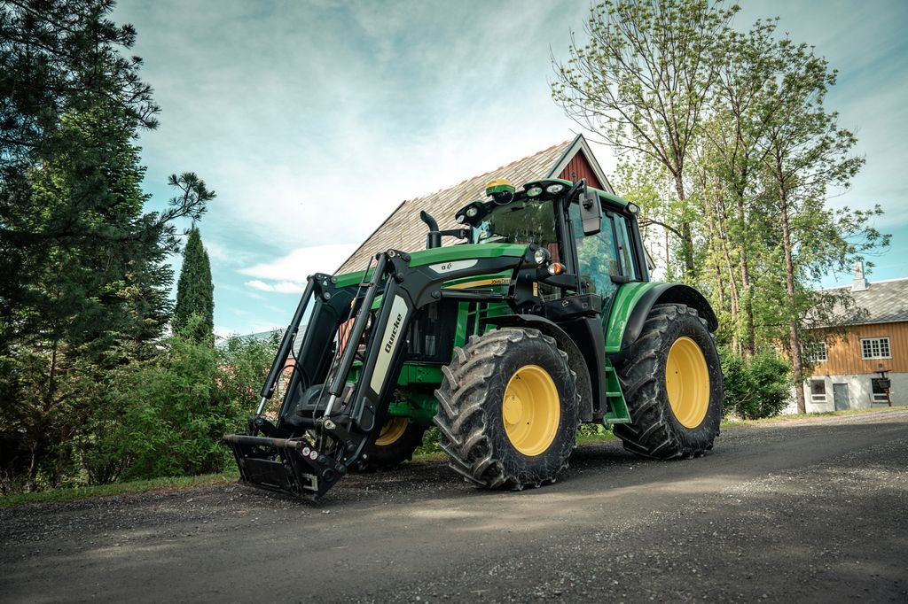 Grønn traktor med lasteapparat står parkert på et gårdstun. Foto.