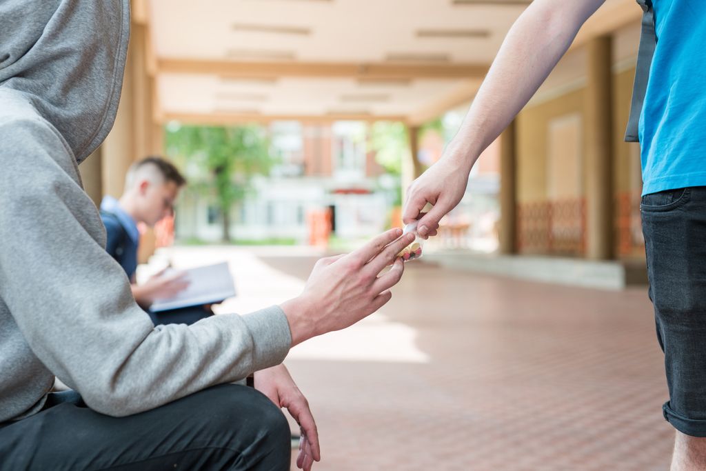 Anonym person i hettegenser mottar en liten pose med fargerike piller eller drops av en forbipasserende. Foto.