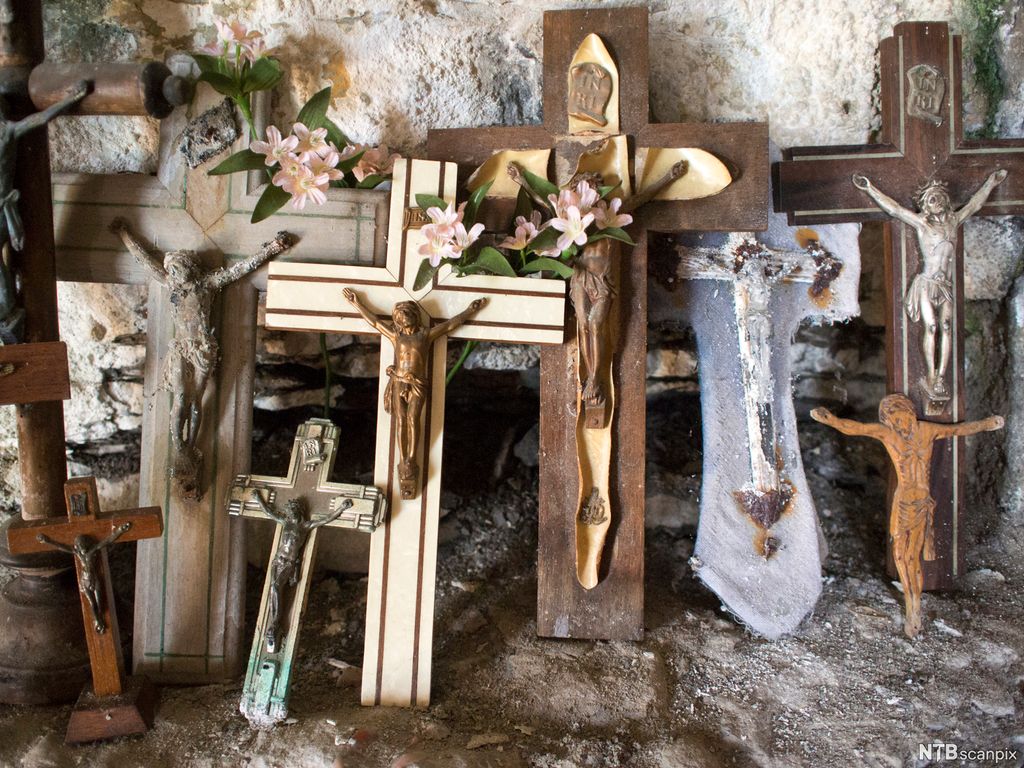 Syv ulike figurer av Jesus på korset stilt opp mot en vegg. De er pyntet med blomster. Foto.