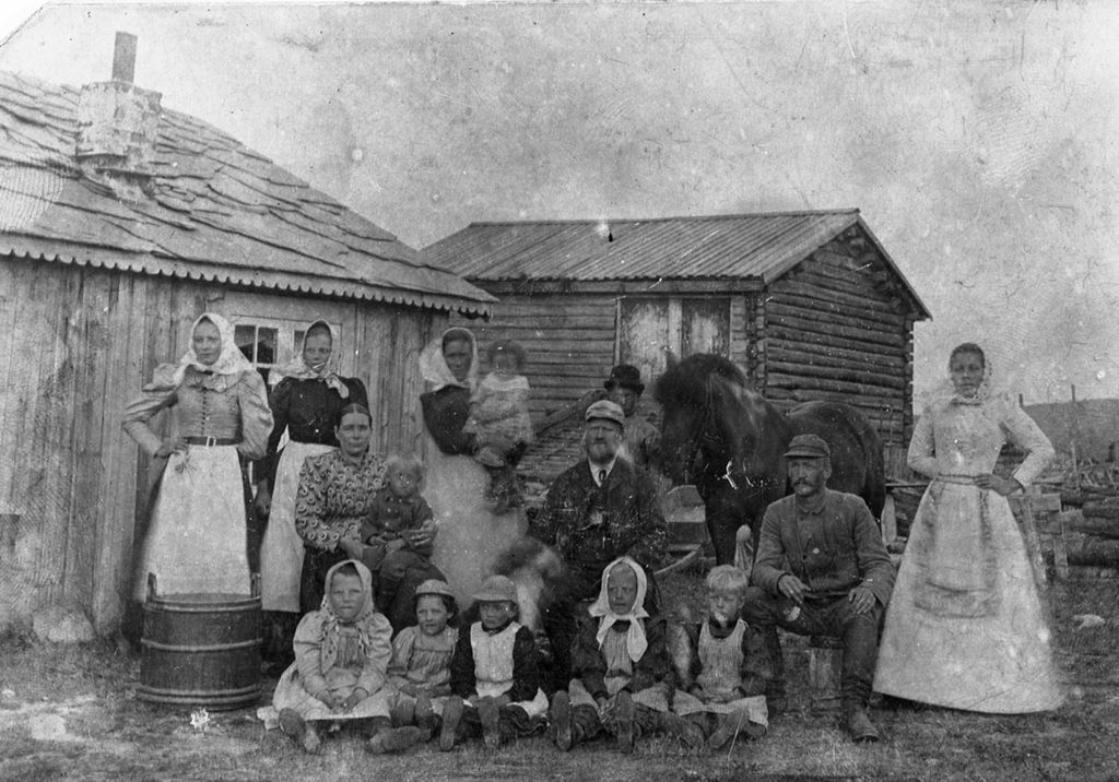 Storfamilie i Stor-Elvdal rundt 1890. Det er flere voksne kvinner og menn pluss mange barn. I bakgrunnen er det små trebygninger og dessuten en hest. Svart-hvitt-foto.