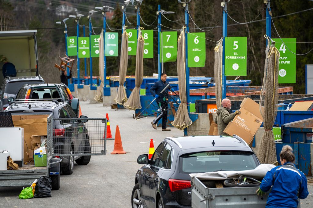 Biler med tilhengere står på rekke og rad mens folk sorterer søppel i diverse konteinere ved en mottaksstasjon. Foto. 