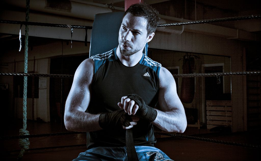 Mann inne i et halvmørkt treningsrom. Han sitter i hjørnet på en boksering og surrer tøystykker rundt hendene sine mens han stirrer alvorlig ut i luften. Foto. 