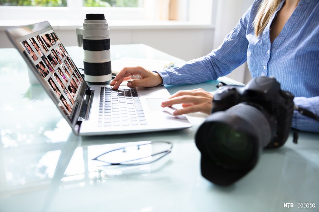 Kvinne jobber på en laptop. Ved siden av laptopen ligger et speilreflekskamera og et stort objektiv. Foto. 
