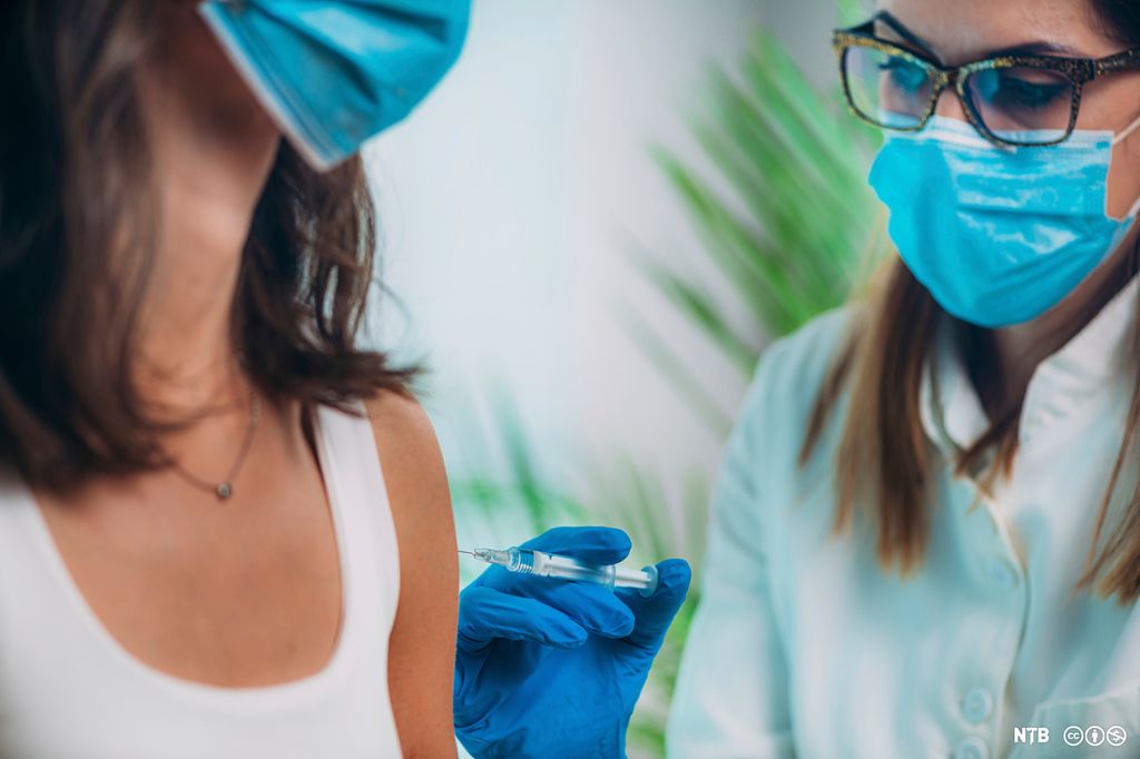 En hvitkledd helsearbeider vaksinerer en kvinne. Begge har munnbind. Foto.