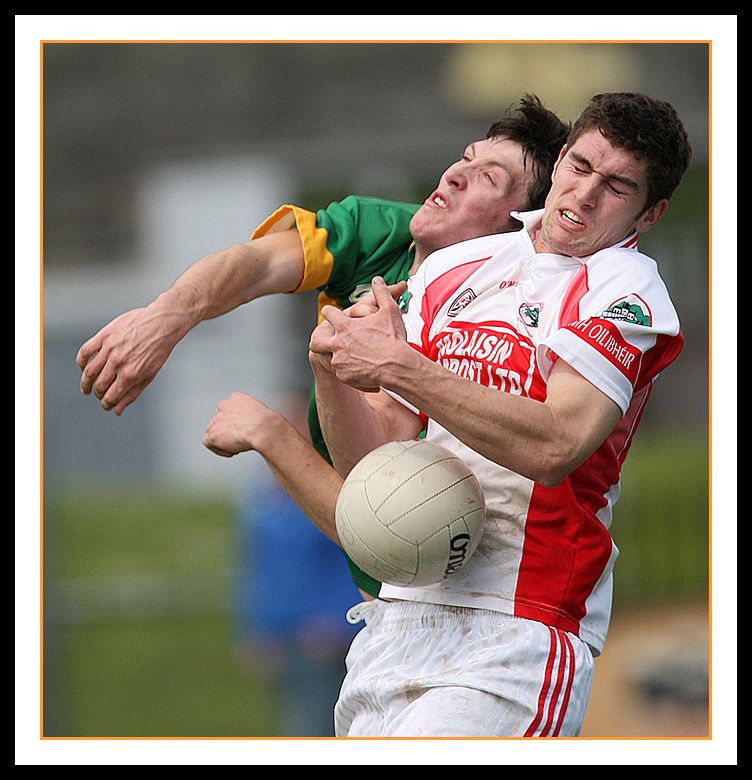 Two players in football kit, one green and yellow, the other white and red, knock against each other while a ball bounces in front of them. Photo.