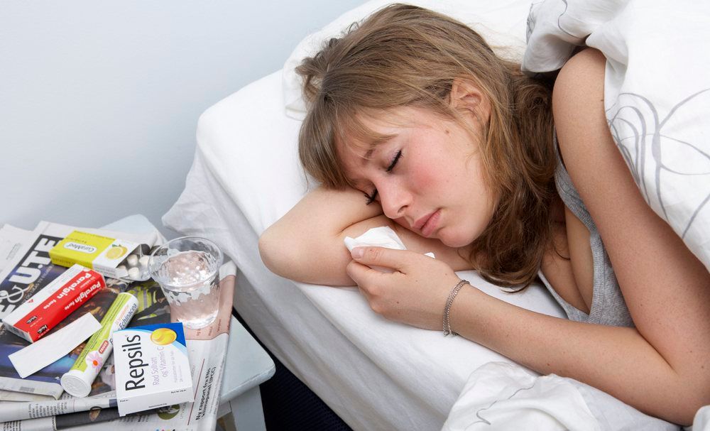 Sjuk ung kvinne ligg i senga og har halstablettar, smertestillande medikament, solhattablettar og vassglas på nattbordet. Foto.