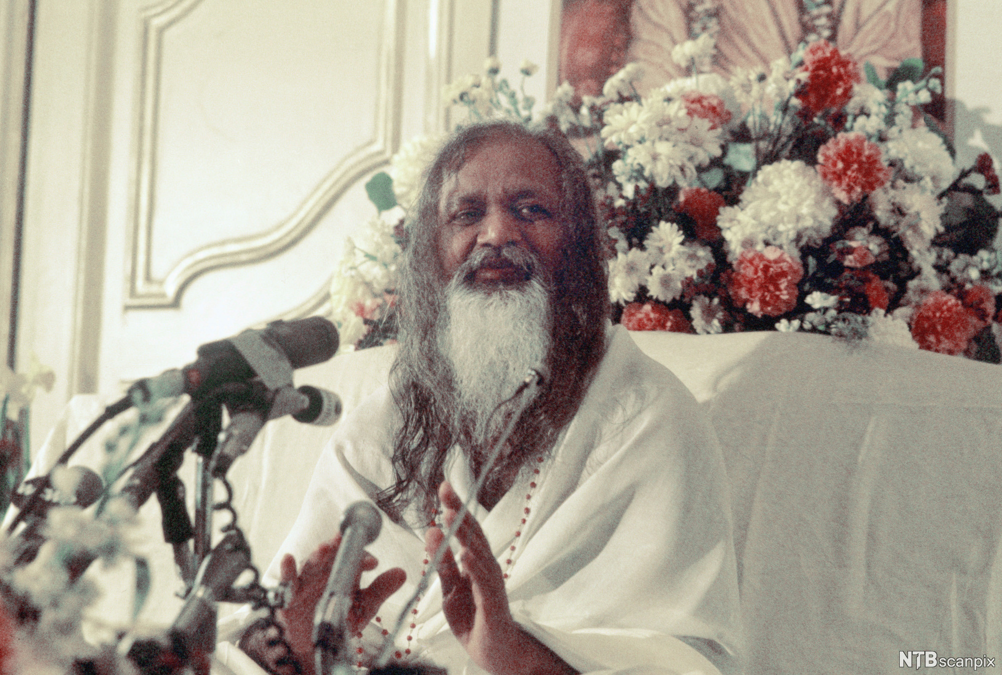 Mann med skjegg og langt hår, kledd i hvitt, står og taler med flere mikrofoner foran seg. Foto.