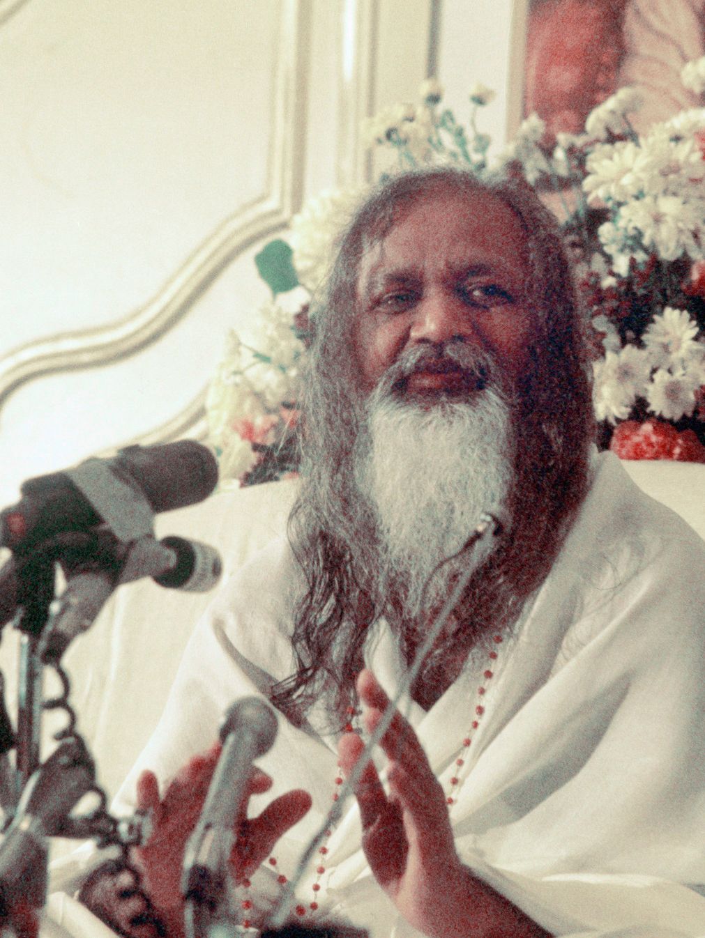 Mann med skjegg og langt hår, kledd i kvitt, står og taler med fleire mikrofonar framfor seg. Foto.