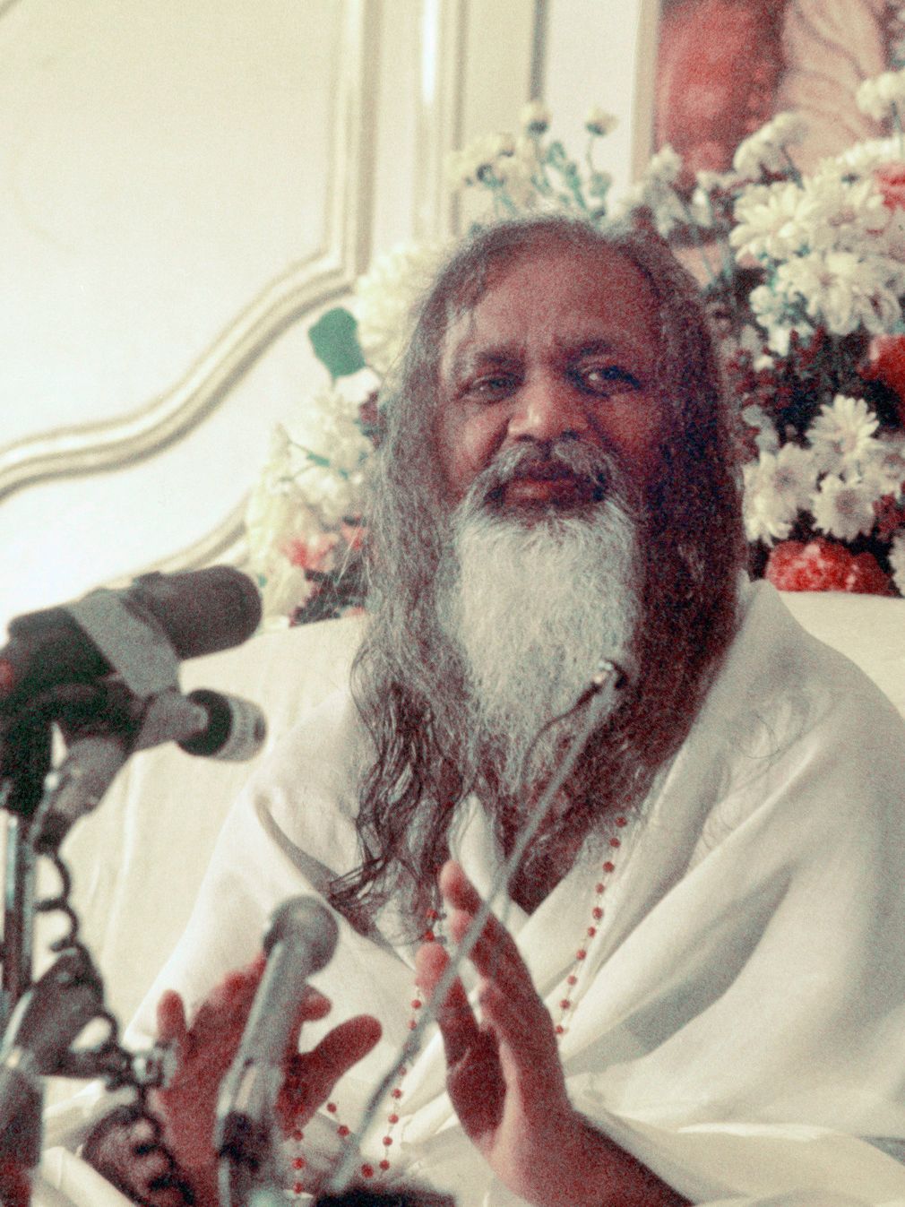 Mann med skjegg og langt hår, kledd i hvitt, står og taler med flere mikrofoner foran seg. Foto.