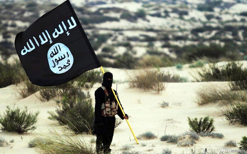 En tildekket mann med våpenbelte og IS-flagg et sted i ørkenen i Irak eller Syria. Foto. 