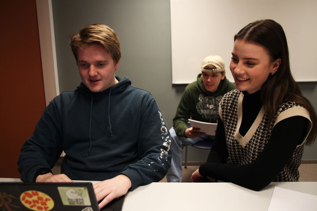 Gutt og jente ser på dataskjerm som gutten bruker. I bakgrunnen sitter en annen gutt med notatblokk og følger med på skjermen. Foto. 