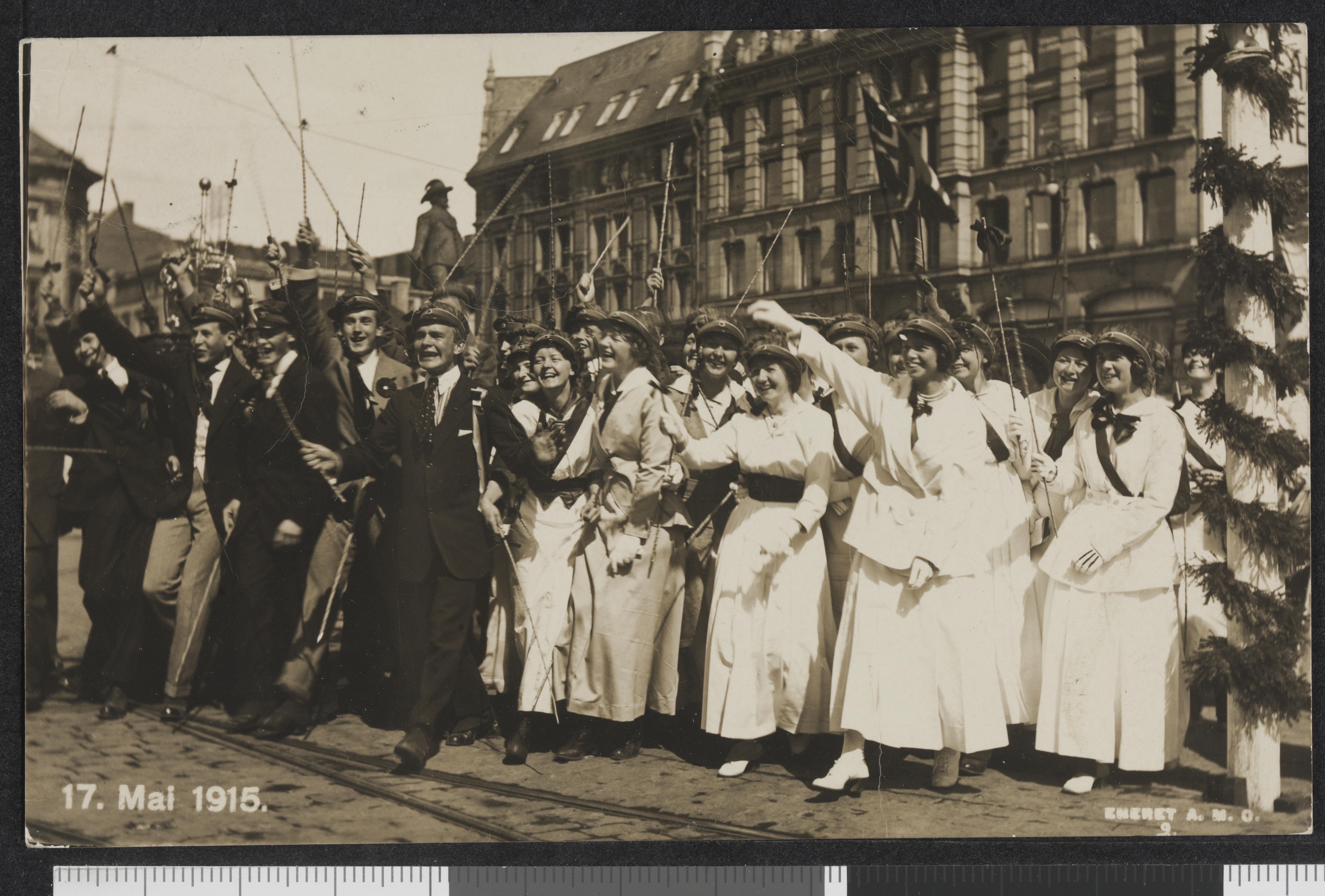 Smilende unge menn i svart dress og unge kvinner i hvite kjoler har russelue på og vifter med bambusstokker. Svart/hvitt-foto.