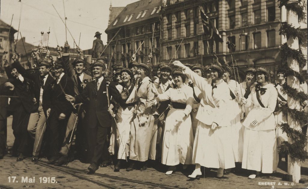 Smilende unge menn i svart dress og unge kvinner i hvite kjoler har russelue på og vifter med bambusstokker. Svart/hvitt-foto.