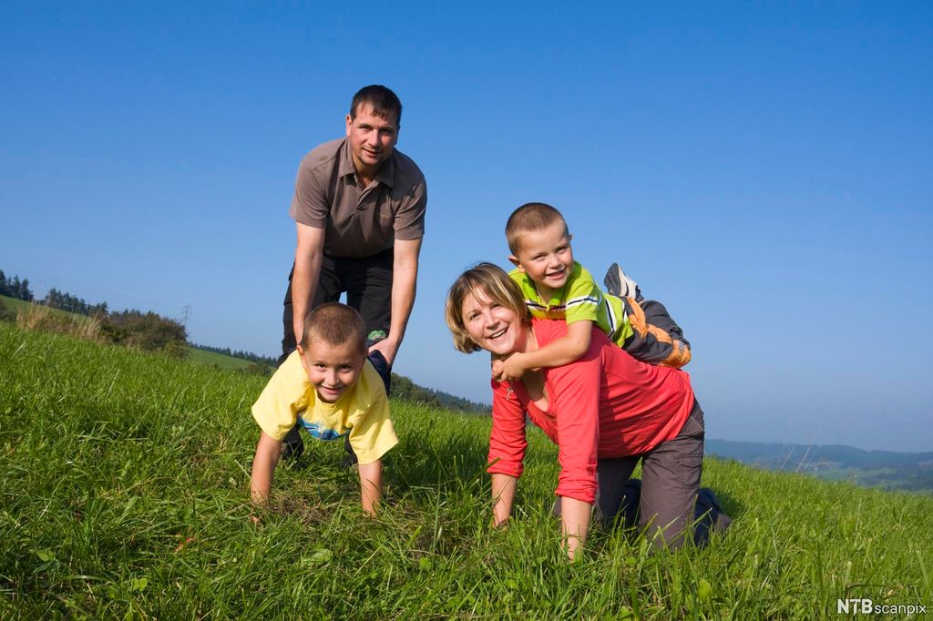 To voksne og to barn leker ute i gresset. En gutt sitter på ryggen til en kvinne som står på alle fire, og en mann holder beina til en annen gutt mens gutten går på hendene. Foto. 