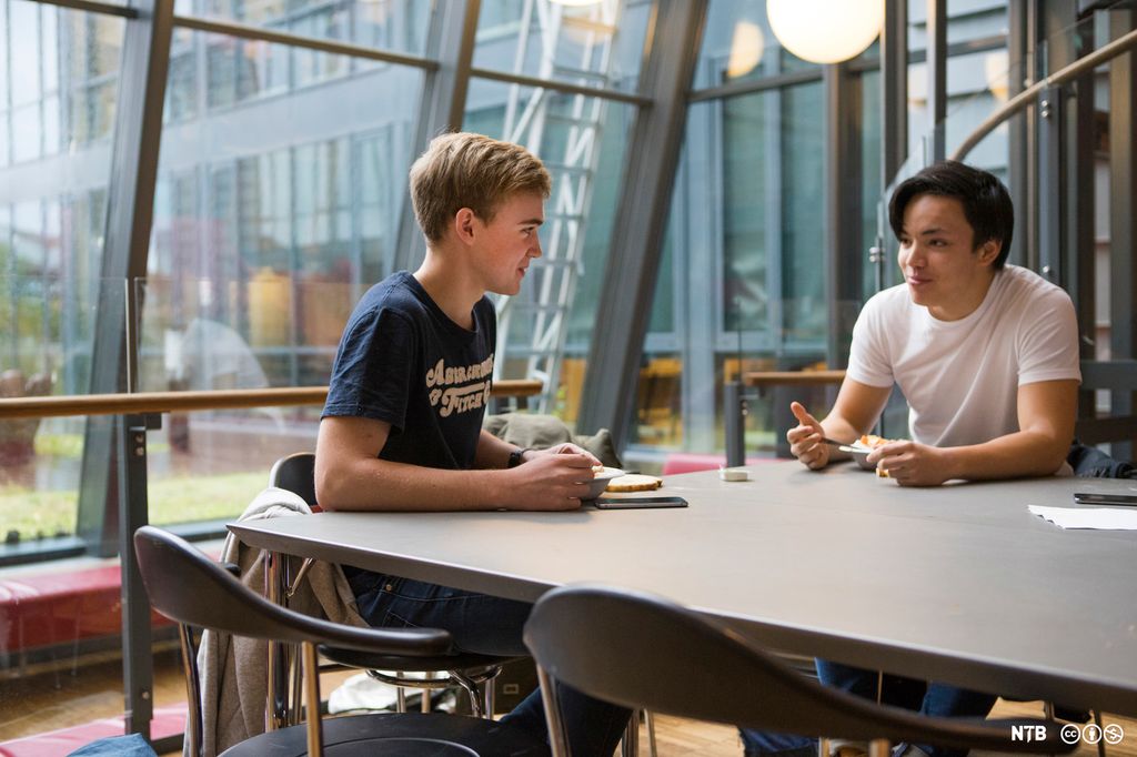 To gutter på videregående skole sitter ved et bord overfor hverandre i kantinen, spiser og snakker sammen. Foto.
