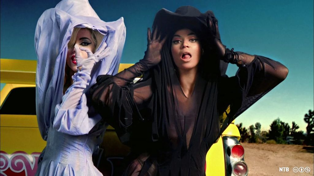 Lady Gaga og Beyoncé står foran en gul bil og gjør grimaser til kameraet. De har på seg flagrete kjoler og matchende bredbremmede hatter med luftige slør – Gaga i hvitt og Beoncé i svart. Foto.