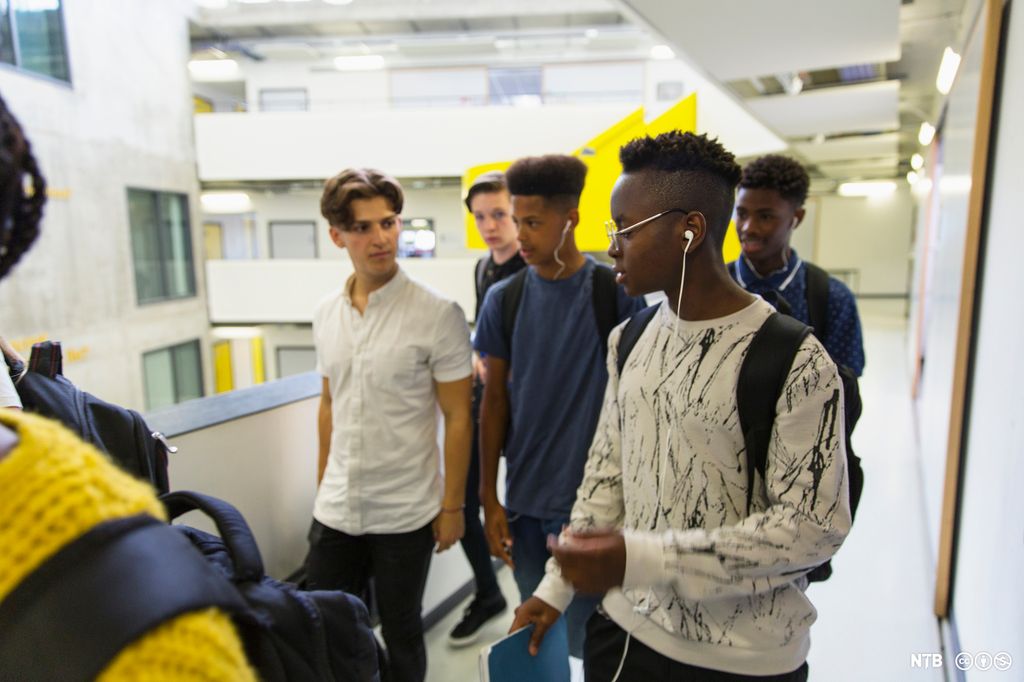 Fem gutter i videregående-alder går sammen i en korridor på en skole. De har sekk på ryggen, den ene gutten holder ei skriveblokk. Foto.